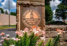 A brown sign with bronze metallic lettering on a stone wall. "A century of excellence" reads atop a bronze crest with the outline of the top of old main. below the image is text reading, "Slippery Rock University 1889-1989" Pink flowers bloom in the sun in front of the placard.